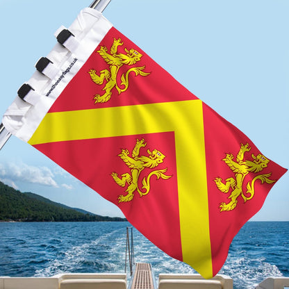 Anglesey county flag with red background and yellow chevron, featuring three gold lions, mounted on a boat rail with a universal triple-lock system, flying at sea with blue sky and coastline in the background