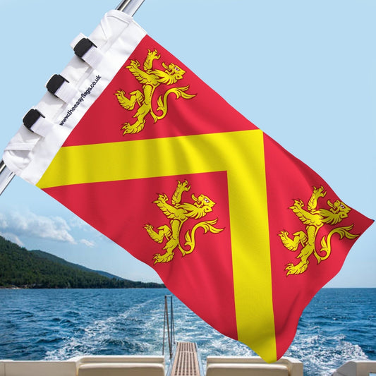 Anglesey county flag with red background and yellow chevron, featuring three gold lions, mounted on a boat rail with a universal triple-lock system, flying at sea with blue sky and coastline in the background