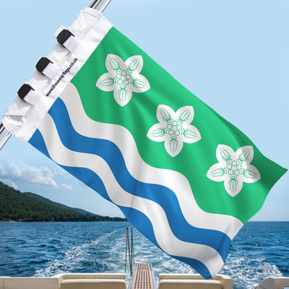 Cumberland county flag with green background, three white parnassus flowers, and blue and white wavy stripes, mounted on a boat rail with a universal triple-lock system, flying at sea with blue sky and coastline in the background