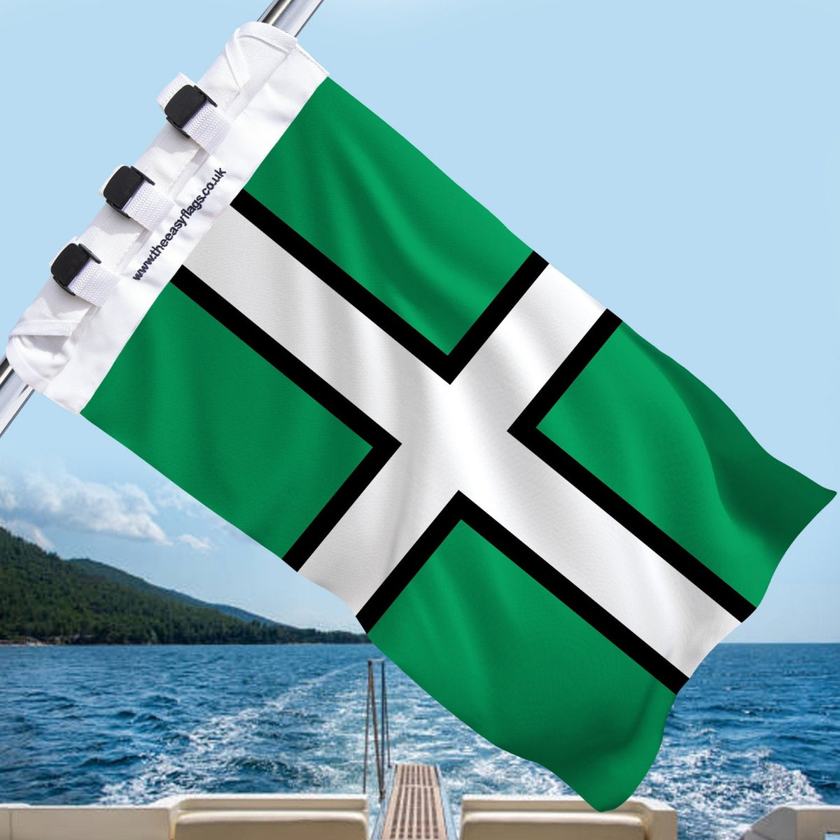 Devon county flag with green field, black cross outlined in white, mounted on a boat rail with a universal triple-lock system, flying at sea with blue sky and coastline in the background