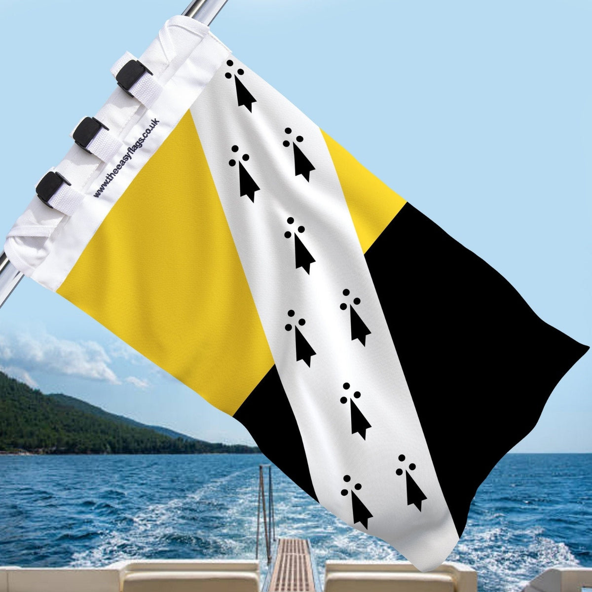 Norfolk county flag with diagonal gold and black field, white bend featuring black ermine spots, mounted on a boat rail with a universal triple-lock system, flying at sea with blue sky and coastline in the background