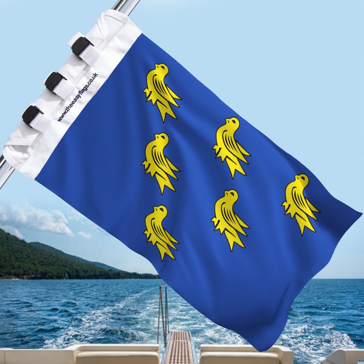 Sussex county flag with blue background and six gold martlets (stylised birds), mounted on a boat rail with a universal triple-lock system, flying at sea with blue sky and coastline in the background